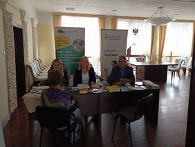 Pokaż zdjęcie: Na zdjęciu dwie kobiety i jeden mężczyzna siedzą przy stole, rozmawiając z kobietą siedzącą naprzeciwko. W tle widoczne są banery Państwowego Urzędu Pracy w Białymstoku i Państwowego Funduszu Rehabilitacji Osób Niepełnosprawnych. Na stole leżą materiały informacyjne.