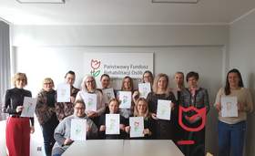 Grupa kilkunastu osób w dwóch rzędach trzyma białe kartki z kolorowymi tulipanami wykonanymi przez uczestników WTZ w Zbąszyniu. W tle, na szarej ścianie, widnieje napis „Państwowy Fundusz Rehabilitacji Osób Niepełnosprawnych” oraz logo PFRON.