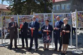 Jasny słoneczny dzień, na rynku na tle wystawy ze zdjęciami prac stoi 6 osób elegancko ubranych, 2 kobiety i czterech mężczyzn, za nimi widać inne osoby