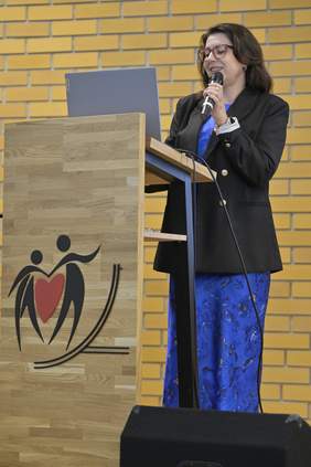 Pokaż zdjęcie: Kobieta w granatowej sukience i czarnej marynarce stoi na scenie, trzymając mikrofon i prowadząc prezentację. Na mównicy widoczny jest symbol przedstawiający postacie trzymające serce. Całość rozgrywa się na tle żółtej, ceglanej ściany