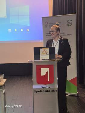 Pokaż zdjęcie: Przy mównicy z napisem gmina Opole Lubelskie stoi kobieta w garniturze.