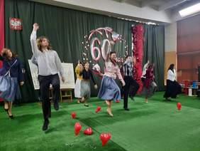 Pokaż zdjęcie: Na środku sali uczniowie wykonują układ taneczny. Podskakują i wyciągają ręce w górę. Obok nich leżą czerwone baloniki w kształcie serduszek. 