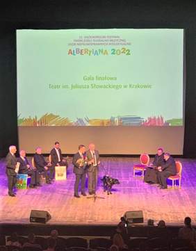Pokaż zdjęcie: Scena teatru. Na scenie siedzą Laureaci. Za nimi wyświetlony napis Albertiana 2022