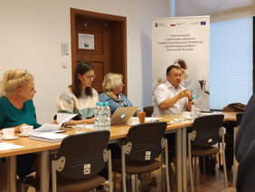 Pokaż zdjęcie: Stół, za nim mężczyzna po prawej stronie, po lwej siedzią trzy kobiety