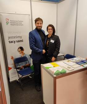 Pokaż zdjęcie: Kobieta i mężczyzna przedstawicieli PFRON stoją przy stoisku informacyjnym podczas Targów Pracy w Białymstoku. W tle widoczny jest baner z hasłem „Wszyscy jesteśmy tacy sami” oraz logo PFRON. Na stoliku znajdują się materiały informacyjne i promocyjne, w tym ulotki oraz broszury dotyczące wsparcia dla osób z niepełnosprawnościami.