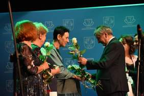 Pokaż zdjęcie: Mężczyzna w garniturze wręcza białe róże trzem kobietom w sukienkach