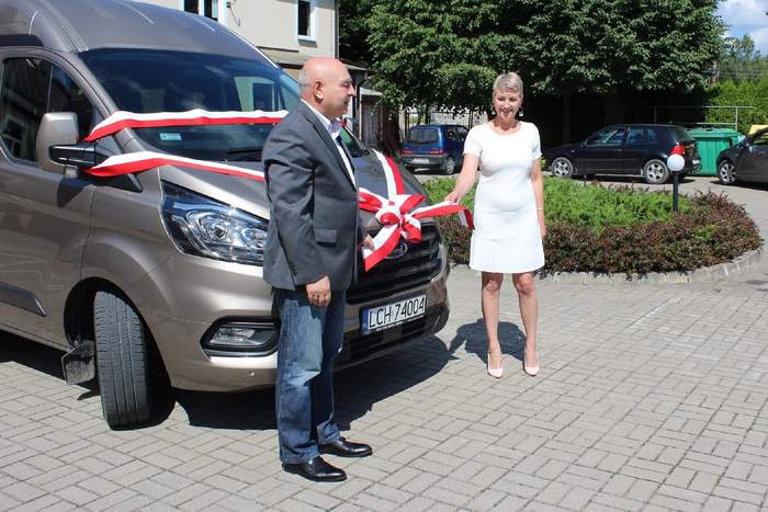 Pokaż zdjęcie: Przed autem na którym jest zawiązana  biało czerwona kokarda stoi mężczyzna w szarej marynarce i trzyma za róg kokardy. Za dugą część kokardy trzyma kobieta ubrana w białą sukienkę. W tle budynek Urzędu, klomb z roślinami.