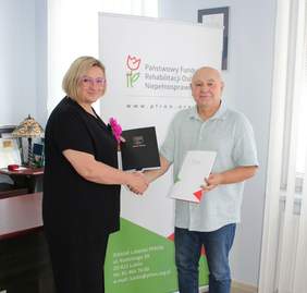 Pokaż zdjęcie: Kobieta blondynka w różowych okularach i czarnym kostiumie pozuje do zdjęcia z mężczyzną ubranym w jasną koszulę. Osoby te ściskają sobie ręce oraz trzymają podpisane umowy. W tle rollup z logo funduszu.