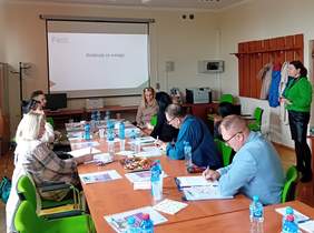 Pokaż zdjęcie: Po prawej i lewej stronie długiego stołu siedzą uczestnicy spotkania, robią notatki. Przed nimi leżą broszury. Na ekranie wyświetla się slajd z napisem Dziękuję za uwagę i logo PFRON. Po prawej stronie sali stoi kobieta w zielonym sweterku.