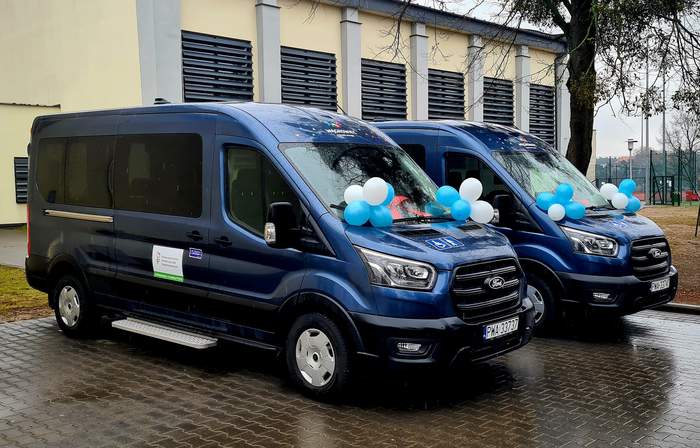 Pokaż zdjęcie: Dwa niebieskie, dziewięcioosobowe busy marki Ford zaparkowane obok siebie na mokrej kostce brukowej przed budynkiem szkoły. Pojazdy ozdobione są biało-niebieskimi balonami. W tle widoczny jasny budynek z ciemnymi żaluzjami oraz drzewo po prawej stronie.