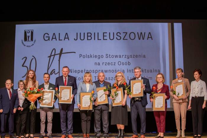 Pokaż zdjęcie: Na scenie podczas 30 lecia gali pozuje 13 osób trzymając w ręku pamiątkowe dyplomy oraz kwiaty
