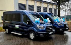 Dwa niebieskie, dziewięcioosobowe busy marki Ford zaparkowane obok siebie na mokrej kostce brukowej przed budynkiem szkoły. Pojazdy ozdobione są biało-niebieskimi balonami. W tle widoczny jasny budynek z ciemnymi żaluzjami oraz drzewo po prawej stronie.