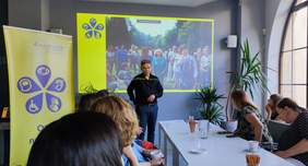 Pokaż zdjęcie: Sala konferencyjna. Mężczyzna na środku sali przemawia do osób siedzących przy stolikach. Za nim, na scianie wyświetlane są zdjęcia. Uczestnicy spotkania słuchają z zainteresowaniem. 