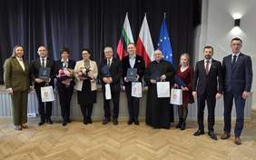 Pokaż zdjęcie: Zdjęcie grupowe ludzi stojących w jednym rzędzie na sali, na tle ciemnej kurtyny i trzech flag. Jest około dziesięciu osób, kobiety i mężczyźni, ubrani głównie formalnie (garnitury, sukienki; jedna osoba w czarnej szacie). Kilka osób trzyma niebieskie teczki, a część osób trzyma białe torby z czerwonym herbem; dwie osoby trzymają bukiety kwiatów. W tle stoją trzy flagi na masztach: biało zielono czerwona, biało czerwona (flaga Polski) oraz niebieska z żółtymi gwiazdami (flaga Unii Europejskiej).