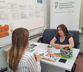 Pokaż zdjęcie: Przy stoliku na którym leżą ulotki, katalogi rozmawiają dwie kobiety. W tle baner z logo PFRON.