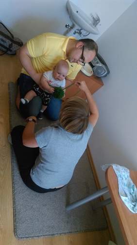 Pokaż zdjęcie: Kobieta z pomponem w ręku siedząca na dywaniku naprzeciwko mężczyzny, który trzyma dziecko na kolanach