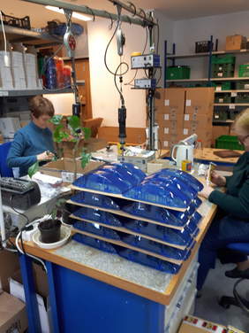 Pokaż zdjęcie: Dwie osoby pracują przy produkcji sygnalizatorów, na pierwszym planie gotowe obudowy, w tle regały magazynowe