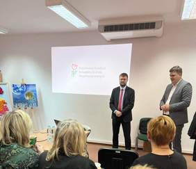 Pokaż zdjęcie: Dwóch mężczyzn w garniturach z uśmiechem na twarzy, przed nimi trzy kobiety