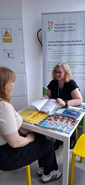 Pokaż zdjęcie: Przy stoliku siedzą dwie kobiety. Jedna z pań szeroko się uśmiecha. Na stoliku leżą różne informatory, poradniki, czasopisma, broszury na temat dofinansowań dla osób z niepełnosprawnością. 