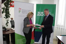 Pokaż zdjęcie: Kobieta i mężczyzna stojący na tle rollupów z logotypami PFRON oraz ZUS wymianiaja się podpisanymi dokumentami