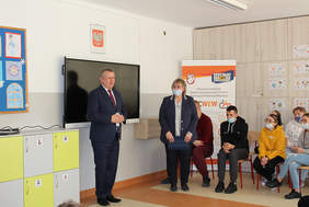 Pokaż zdjęcie: Przed tablicą multimedialną stoi mężczyzna w garniturze, przemawia, obok kobieta w okularach i maseczce w tle dzieci siedzące na krzesełkach