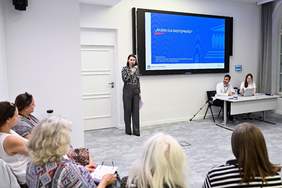 Pokaż zdjęcie: Na pierwszym planie, po lewej stronie, siedzi grupa słuchaczy. Po prawej stronie, przy stole, siedzi dwóch prowadzących z laptopami. Po środku stoi kobieta trzymająca mikrofon, przemawiająca do publiczności. Na ścianie za nią znajduje się duży ekran z napisem "RAZEM DLA DOSTĘPNOŚCI". Na ekranie widoczne są także logotypy PFRON oraz Biura Rzecznika Praw Obywatelskich.