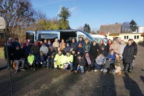 Pokaż zdjęcie: Kilkadziesiąt osób stoi na zewnątrz. Ubrani są w kurtki. Za nimi granatowy mikrobus z otwartymi drzwiami. W tle drzewa i budynki