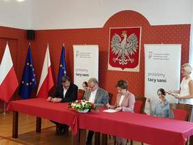 Pokaż zdjęcie: Trzy osoby siedzą przy stole konferencyjnym, dwie podpisują dokumenty.