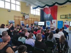 Pokaż zdjęcie: Kilkadziesiąt osób siedzi na krzesłach w dużej sali. Przed nimi kobieta z mikrofonem oraz liczne banery promujące instytucje