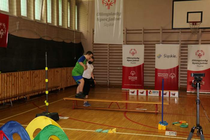 Pokaż zdjęcie: Fragment sali gimnastycznej na długiej, niskiej ławce stoi chłopak w zielonogranatowym stroju i próbuje po niej przejść jest asekurowany przez instruktorkę