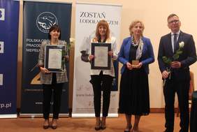 Pokaż zdjęcie: Na scenie stoją trzy kobiety i mężczyzna. Jedna z kobiet ma w ręku białą różę oraz dyplom w ram kach.Druga trzyma dyplom,trzecia statuetkę Lodołamacza. Mężczyzna w okularach i garniturze trzyma białą róże  