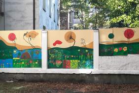 Pokaż zdjęcie: Mur pomalowany przez dzieci i osoby z niepełnosprawnościami. Obrazy na murze to kolorowe kwiaty otoczone zielenią, błękitne niebo i drzewa. Za murem widoczny fragment starej kamienicy.