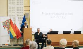 Pokaż zdjęcie: Na zdjęciu Edyta Kondzior stoi przy mównicy podczas konferencji, witając gości. Obok niej znajduje się tłumacz języka migowego, a w tle widać ekran z prezentacją i flagi Polski, Unii Europejskiej oraz Województwa Podlaskiego. Publiczność siedzi przodem do prelegentów