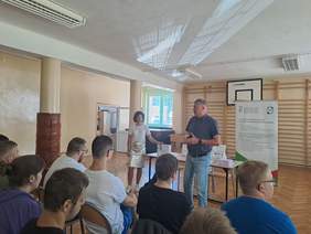 Pokaż zdjęcie: Z tyłu sali w kierunku dwóch prelegentów, młoda kobieta oparta jest o stolik, mężczyzna wchodzi w interakcję ze słuchaczami.