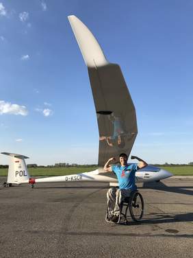 Pokaż zdjęcie: Mężczyzna na wózku pozuje z uniesionymi i zgiętymi ramionami w geście siły na tle szybowca stojącego na płycie lotniska. Ubrany jest w niebieską koszulkę i czapkę z daszkiem. Widać jedno skrzydło szybowca. W tle zielone pole i niebo z lekkimi chmurami