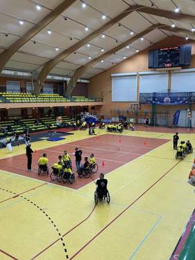 Pokaż zdjęcie: Widok wnętrza dużej hali sportowej. Na boisku oznaczonym liniami znajduje się kilka osób poruszających się na wózkach. W tle widoczne są trybuny, tablica wyników oraz elementy wyposażenia hali.)