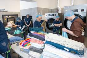 Pokaż zdjęcie: Osoby pracujące w pralni przemysłowej sortują, składają i przygotowują wyprane tekstylia. W tle widoczne są duże przemysłowe pralki oraz kosze z praniem.