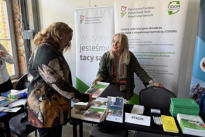 Pokaż zdjęcie: Pracowniczka stoiska informacyjnego CIDON przekazuje materiały informacyjne odwiedzającej. Na stole znajdują się broszury, ulotki i długopisy, a w tle widoczne są roll-upy PFRON oraz CIDON.