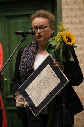 Pokaż zdjęcie: Na scenie przed mikrofonem stoi kobieta ubrana w ciemny kostium, białą bluzkę a na szyi przewieszony ma ciemny szal z niebiesko szarymi wzorami. Na nosie nosi okulary. W prawej dłoni trzyma statuetkę lodołamaczy, w lewej trzyma kwiaty a dyplom trzym