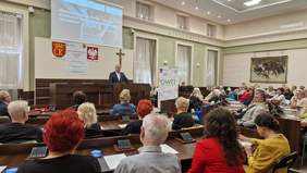 Pokaż zdjęcie: mężczyzna stoi na mównicy, przed nim słuchacze zasiadający na sali obrad w Urzędzie Miasta w Kielcach