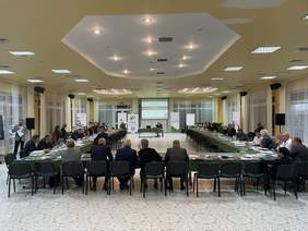 Pokaż zdjęcie: Na dużej jasno oświetlonej sali stoi stół w kształcie litery U. Po zewnętrznej stronie siedzą na galowo ubrani ludzie. Na końcu sali wisi ekran z wyświetloną prezentacją.