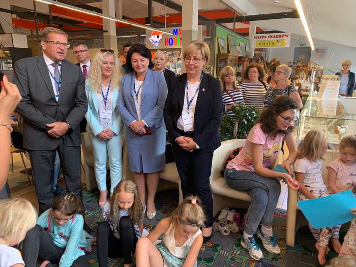 Pokaż zdjęcie: We wnętrzu galerii cztery osoby w eleganckich strojach stoją wśród dzieci malujacych obrazki. Dzieci siedzą na podłodze. Z tyłu wiele postaci przyglądajacych się.