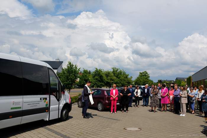 Pokaż zdjęcie: Duża grupa osób, w tym między innymi ksiądz, stoi na placu wyłożonym kostką brukową. Z przodu widoczny jest biały autobus z logotypem Warsztatu Terapii Zajęciowej w Tarnowie Podgórnym oraz naklejką informującą, że pojazd dostosowany został do przewozu osób poruszających się na wózku. W tle widoczne są drzewa liściaste oraz zabudowania zarówno z płaskim, jak i ze skośnymi dachami. Po lewej stronie możemy dostrzec zaparkowane samochody osobowe. Niebo pokryte jest wypiętrzającymi się chmurami kłębiastymi.
