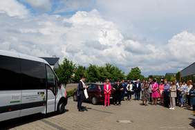 Pokaż zdjęcie: Duża grupa osób, w tym między innymi ksiądz, stoi na placu wyłożonym kostką brukową. Z przodu widoczny jest biały autobus z logotypem Warsztatu Terapii Zajęciowej w Tarnowie Podgórnym oraz naklejką informującą, że pojazd dostosowany został do przewozu osób poruszających się na wózku. W tle widoczne są drzewa liściaste oraz zabudowania zarówno z płaskim, jak i ze skośnymi dachami. Po lewej stronie możemy dostrzec zaparkowane samochody osobowe. Niebo pokryte jest wypiętrzającymi się chmurami kłębiastymi.