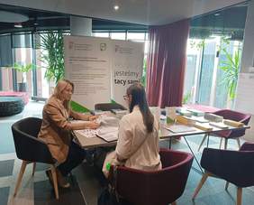 Pokaż zdjęcie: Kobieta siedząc przy stoisku Oddziału Wielkopolskiego PFRON udziela informacji innej kobiecie. Trzymając w ręce ulotkę, opiera się o stolik, na którym rozłożone są materiały informacyjne. W tle banery PFRON i CIDON. Na sali są też kwiaty doniczkowe.