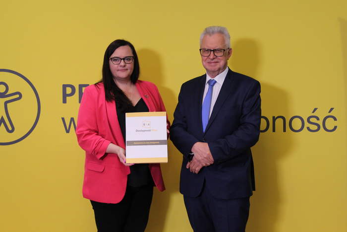 Pokaż zdjęcie: Na tle żółtej ścianki stoi ciemnowłosa kobieta w okularach trzymająca w rękach dokument, obok niej siwowłosy mężczyzna ubrany garnitur. 