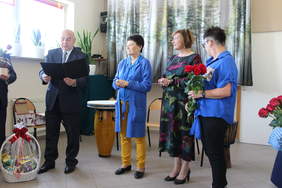 Pokaż zdjęcie: Na głównym planie stoi mężczyzna w garniturze i czyta list obok stoją trzy kobiety