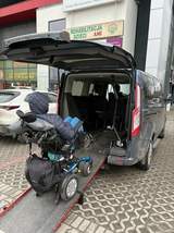 Osoba siedząca na wózku inwalidzkim wjeżdża do wnętrza samochodu przystosowanego do przewozu osób z niepełnosprawnością. Tylna klapa pojazdu jest podniesiona. W tle widać miejskie budynki. 