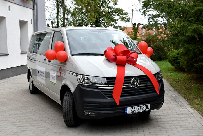 Pokaż zdjęcie: Bialy bus, owinięty czerwoną kokardą, z logo PFRON naklejonym na drzwiach auta, stoi przed budynkiem Szkoły