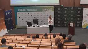 Pokaż zdjęcie: W auli przy stoliku stoi kobieta, skierowana do uczestników spotkania. Zdjęcie robione z auli,  widać rzędy krzeseł, zajęte są pojedyncze miejsca.